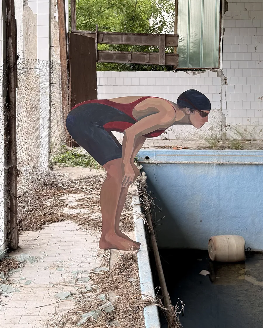 Close view of the painted swimmer leaning over the pool edge, with broken windows and greenery behind.